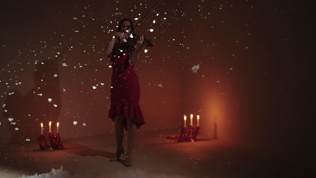 Donna che suona il violino con un vestito rosso, tra fiocchi di neve e candele
