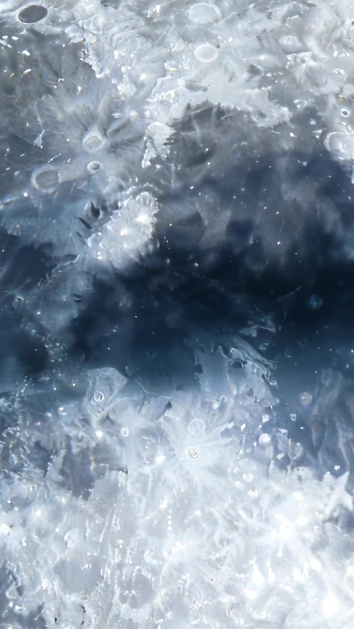 Video verticale: Telecamera che riprende l'acqua dall'alto, con schiuma che si sposta, una banda scura, puntini che galleggiano e bagliori.