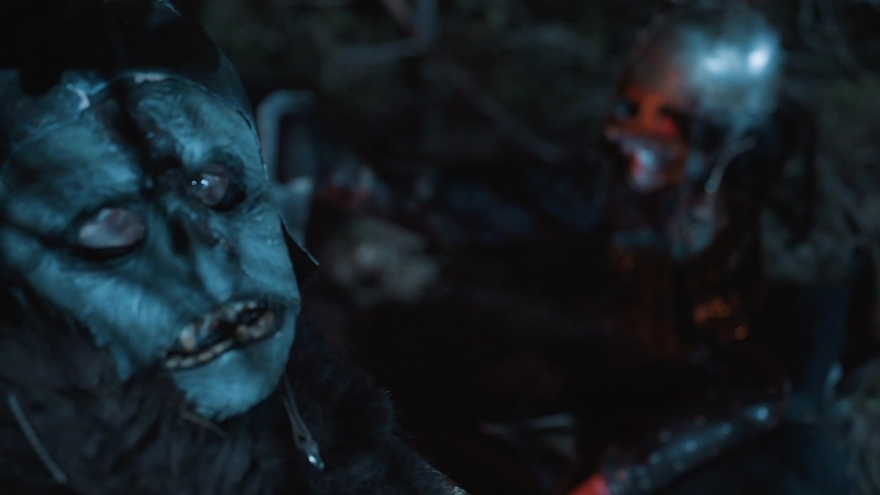 Foresta scura e umida di notte mostra due figure mascherate in un teso incontro ravvicinato, una maschera pallida domina il primo piano mentre una figura con elmetto si accovaccia dietro tra fango, fogliame, ombre, macchie di sangue e un'atmosfera inquietante