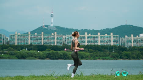 woman in sport clothes doing cordless jump rope exercises jumping on one and both legs at public hangang park with famous n seoul namsan tower and mountain view in background on hot summer day