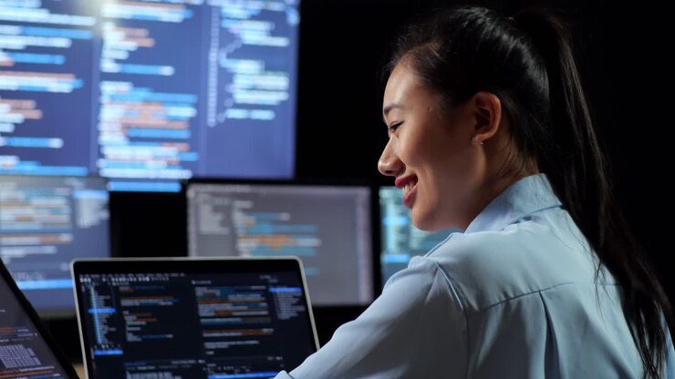 Close Up Back View Of Asian Female Programmer Looking At Database On Tablet And Writing Code By A Laptop Using Multiple Monitors Showing Database On Desktops In The Office