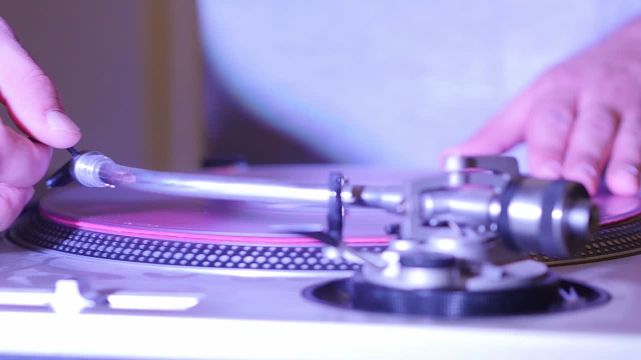 DJ Placing Vinyl On Turn Table And Placing Stylus To Begin Mixing Music While Colored Lights Flash