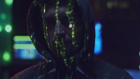 portrait of the young man hacker in a hood looking straight to the camera with projected code numbers and characters on his face in the dark room full of computers