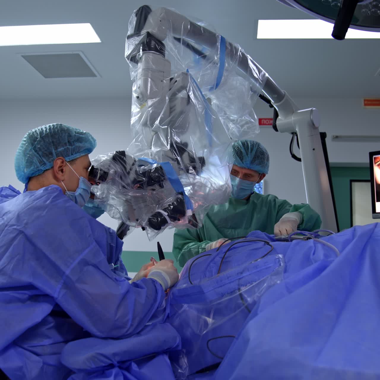 Surgeon sitting near the patient and operating him looking at high-precise microscope. Screen at backdrop shooting the procedure