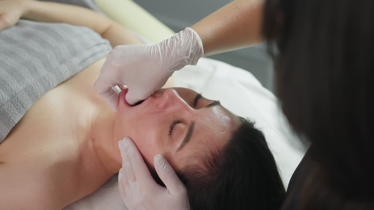 High angle of female client lying on massage chair, closes eyes gently, relaxed as skin care expert wearing surgical gloves dips fingernail inside mouth and uses other hand to massage cheeks in spa