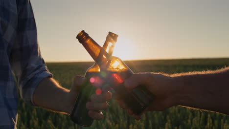 las manos de dos hombres con botellas de cerveza sonando contra el fondo de un campo de trigo donde se pone el sol