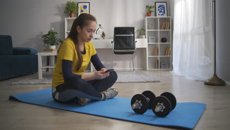 Una-Mujer-Joven-Descansa-Sobre-Una-Alfombra-Deportiva-Con-Pesas-Usando-Un-Teléfono-Inteligente-Para-Navegar-Por-Internet-Haciendo-Ejercicio-En-Casa,-Adicción-A-Las-Redes-Sociales