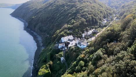 Bucks-Mills-Luftdrohne-Fliegt-Entlang-Der-Bewaldeten-Küste-Von-Devon,-Kleines-Dorf-Auf-Der-Linken-Seite-Des-Rahmens,-Sonniger-Tag,-Strand-Im-Schatten