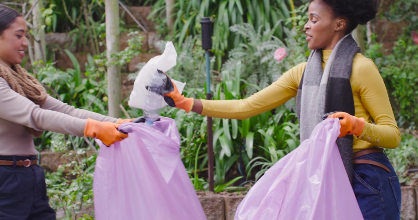 Diverse women with orange gloves spotting and picking bottle, placing in purple bag yard cleanup