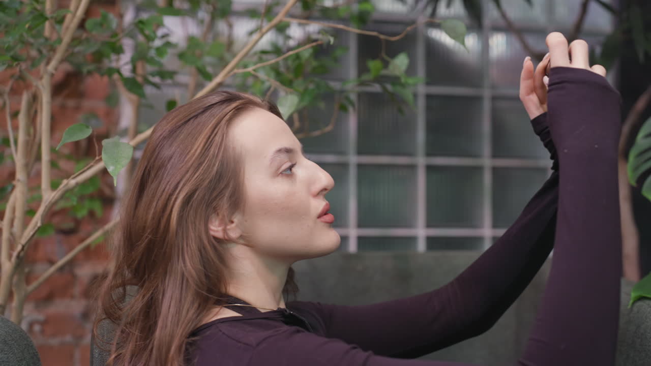 Pretty woman holding phone in landscape orientation, taking picture of green leaf while standing near cushioned chairs with glass panel and brick wall background surrounded by indoor plants