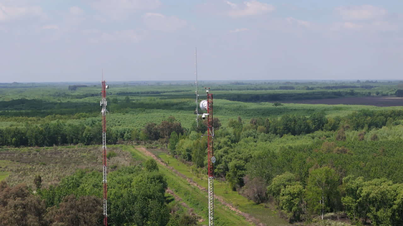 Tall communication tower stands in a rural landscape, surrounded by greenery and open fields, aerial approach