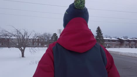 Young-boy-walking-in-snow-on-side-of-road-in-winter-wearing-a-blue-and-red-winter-jacket-and-a-blue-toque-with-a-pompom-on-top