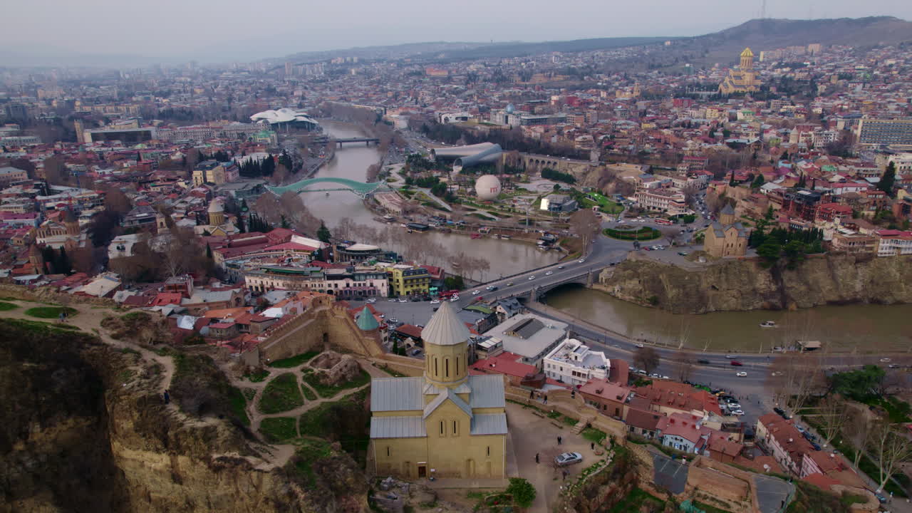 Pohled z dronu na kostel v historickém Tbilisi na útesu, říční mosty, panorama Gruzie, letecká perspektiva cestování, střechy moderních městských čtvrtí a vrstvená městská krajina s parky a současnou architekturou v pozadí.