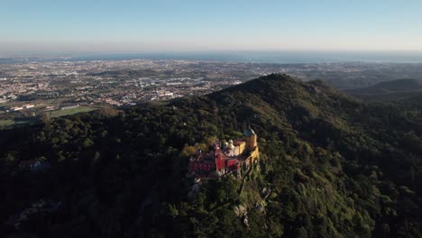 dron dando vueltas alrededor del histórico castillo del palacio pena de pie en la cima de una colina en las montañas de sintra y revelando la ciudad de sintra en el fondo, portugal