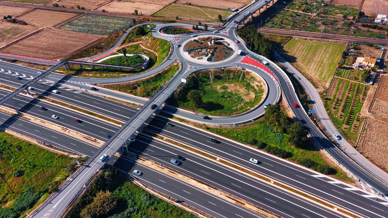 Aerial highway interchange with roundabout and agricultural fields. Aerial view of highway interchange, roundabout, ramps, fields, rail line, and vehicles in daylight