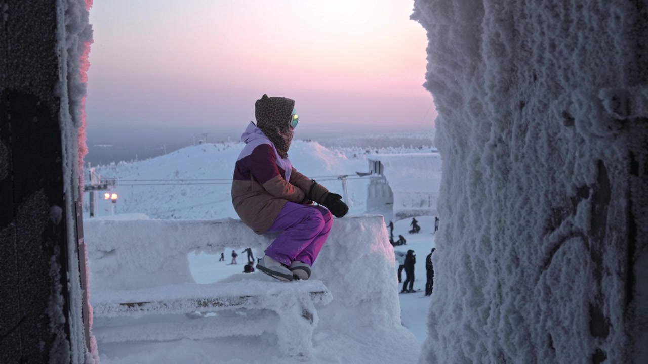 Kayak merkezinde snowboard kıyafetleriyle oturup gün batımını izleyen kişi