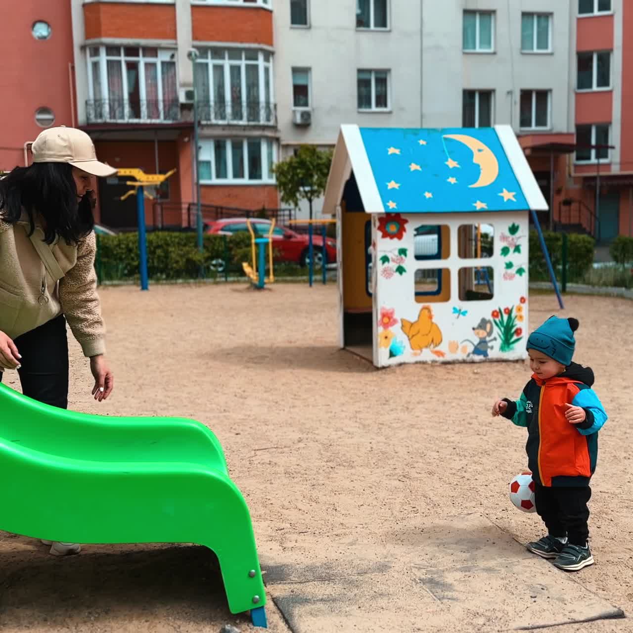 Mother plays with her baby boy on the playground. Woman rolls the ball by the slide and kid runs by the toy