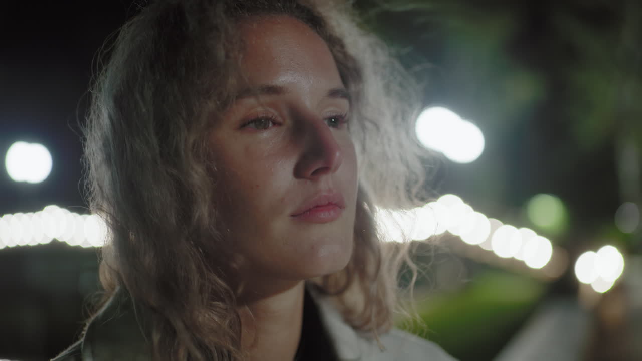 Contemplative woman with light curly hair stares ahead with solemn expression as breeze gently sways her hair, illuminated by glowing bokeh lights and soft reflections in quiet nighttime park