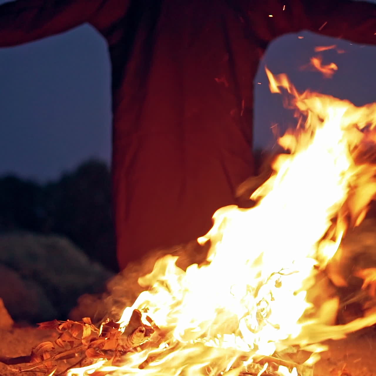 Fire explodes on the ground before silhouette of death. Black robe waving arms near the bright flame in the evening. Horror ghost at night.
