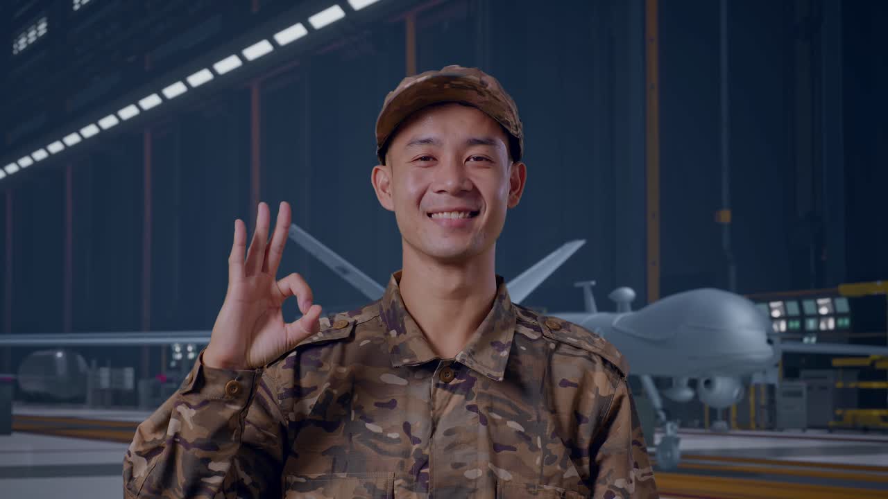 Primo piano di un soldato asiatico che sorride e fa il gesto "OK" alla telecamera mentre è in piedi accanto a un moderno drone militare in un hangar di manutenzione.