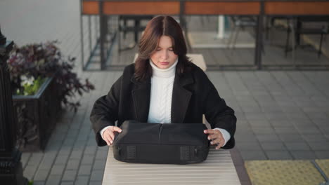 freelancer seated outdoors unzips black laptop bag on a wooden table, background features chairs, tables, and flower vessels, creating a calm urban setting for work