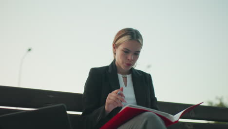 thoughtful white lady carefully studying book while seated alone on wooden bench with laptop beside her in quiet outdoor park setting surrounded by empty benches trees and pole under bright sky