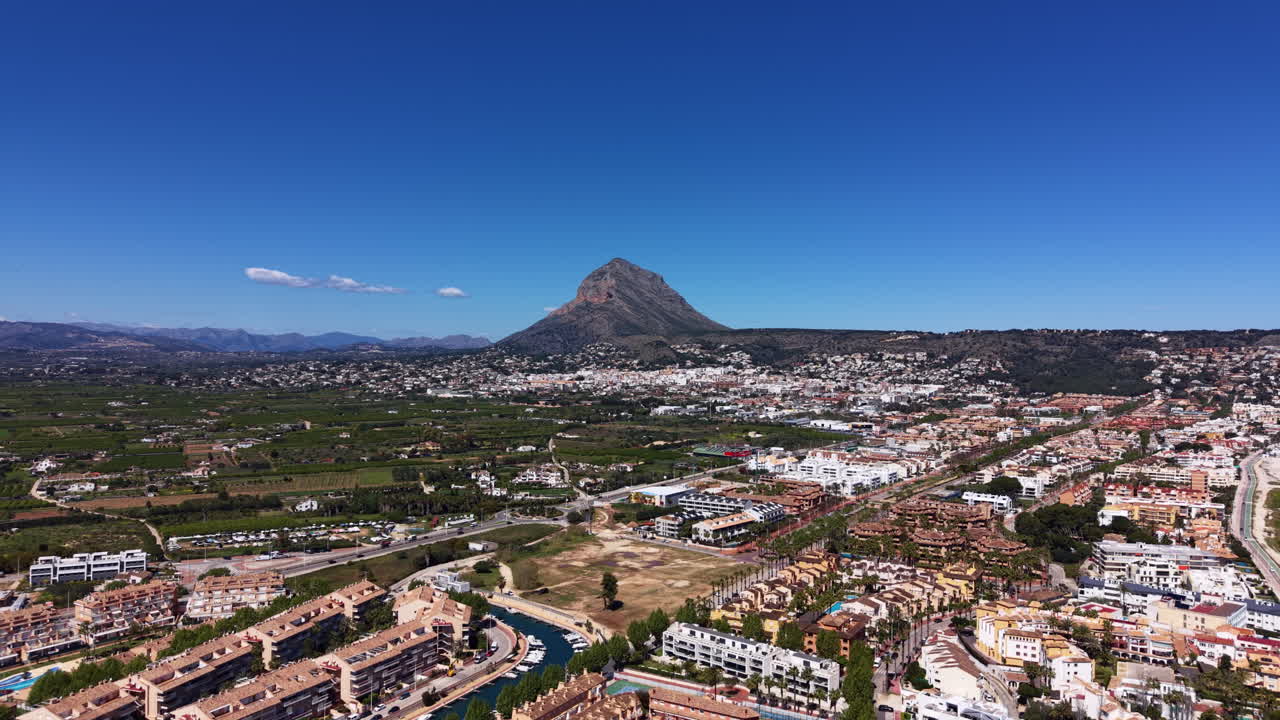 Vista panorâmica de Javea, Espanha, com a montanha Montgó elevando-se acima da cidade costeira, edifícios residenciais, campos verdes e céu azul claro.
