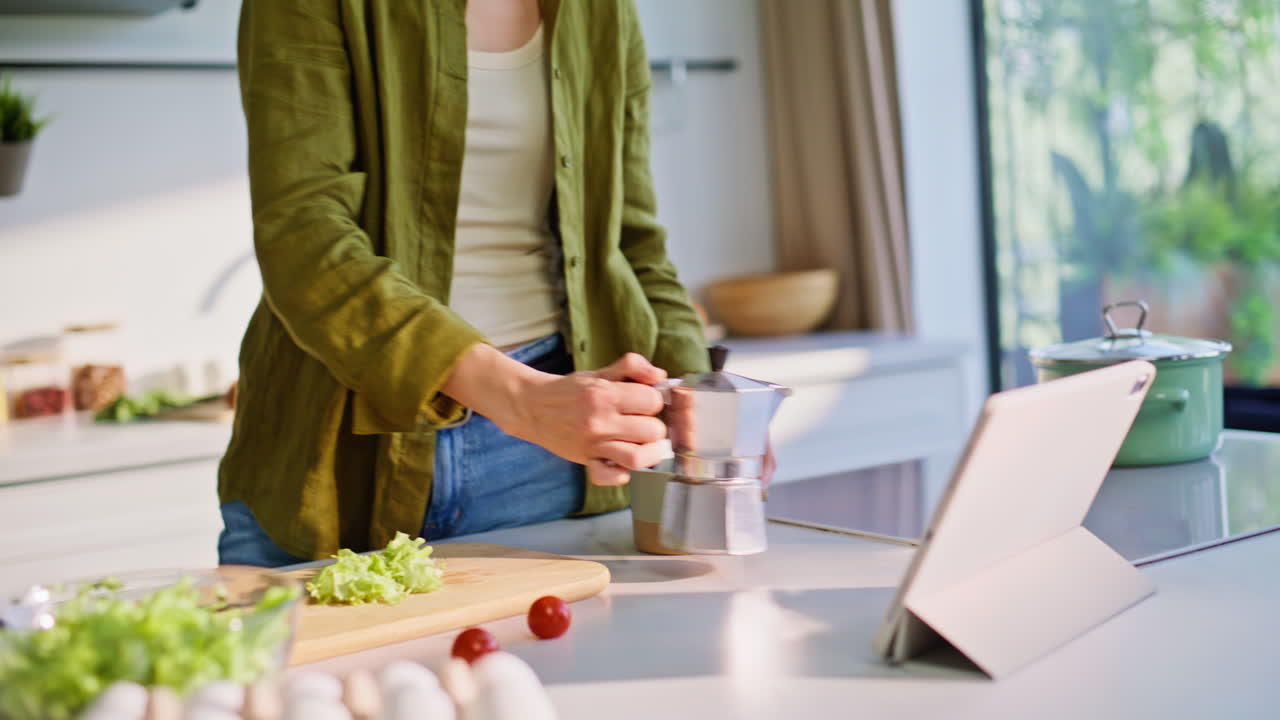 Primo piano di una mano che tocca un tablet mentre guarda un programma mattutino e versa il caffè in cucina