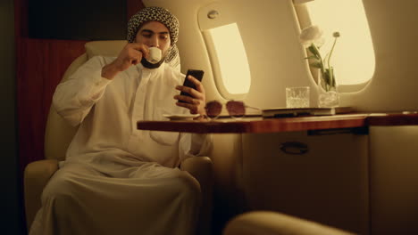 happy man looking cellphone in private jet. smiling arabian rest drinking coffee