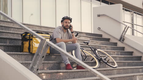 food delivery man on a break seated on a step speaking on the phone next to a thermal backpack