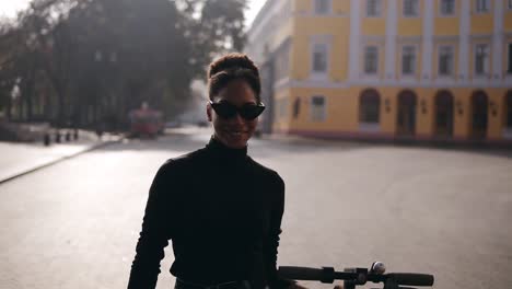 Chica-Afroamericana-Atractiva-Y-Segura-Con-Elegantes-Gafas-De-Sol-Y-Jeans-Azules-Caminando-Despreocupada-Por-La-Calle-De-La-Ciudad-Por-La-Mañana-Con-Su-Scooter-Eléctrico-Negro.-Vista-Frontal.-Camara-Lenta