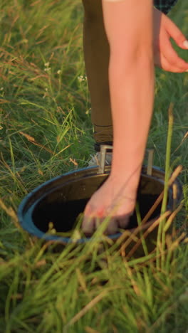 un primer plano de la mano de una mujer apretando un paño, haciendo que el agua gotee en un cubo negro en un campo de hierba cerca de una bicicleta