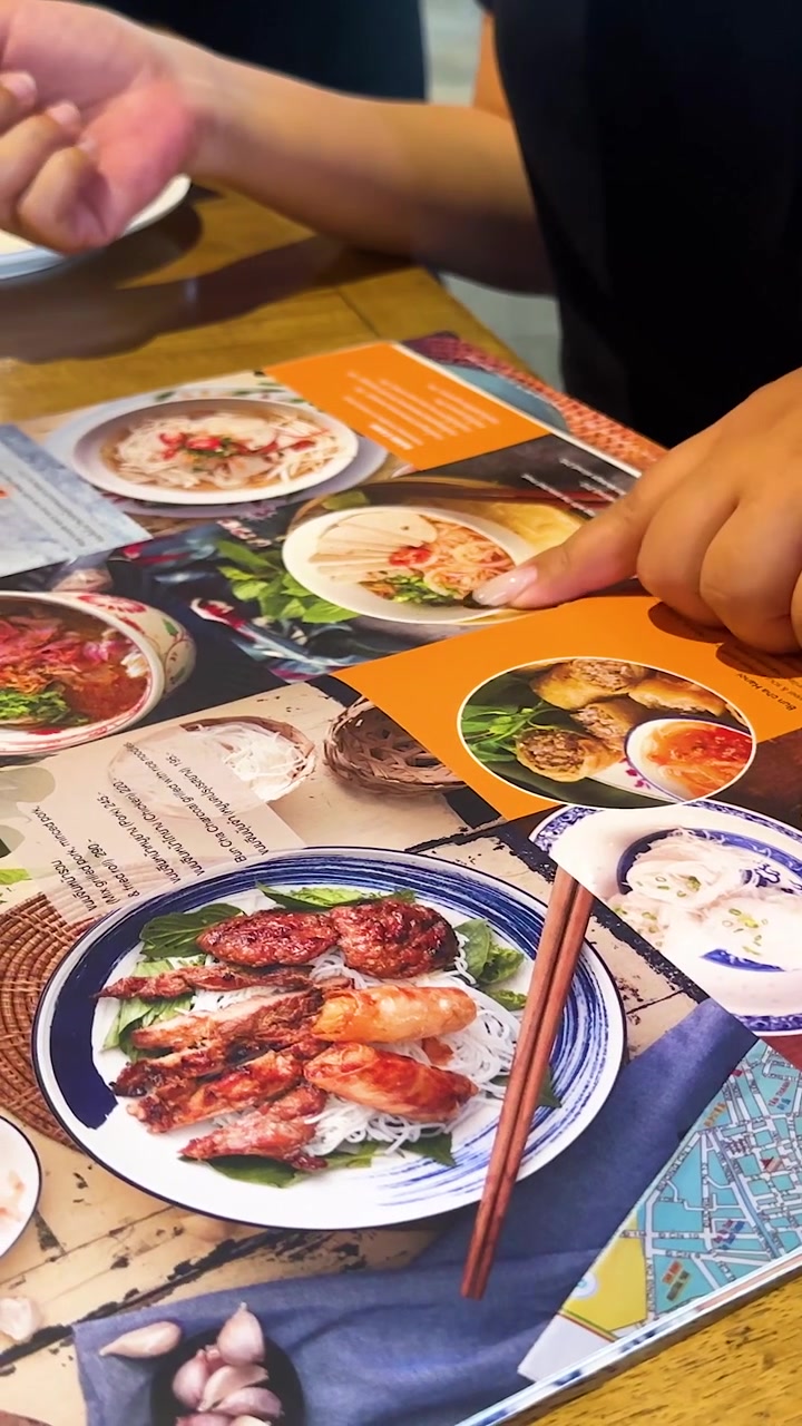 Hender peker på en vietnamesisk meny i en restaurant, som fremhever ulike retter. Sterk belysning og uformell atmosfære.