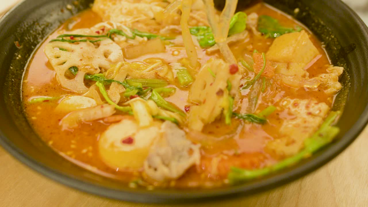 Person lifts noodles and vegetables from hot spicy broth in black bowl, warm indoor lighting