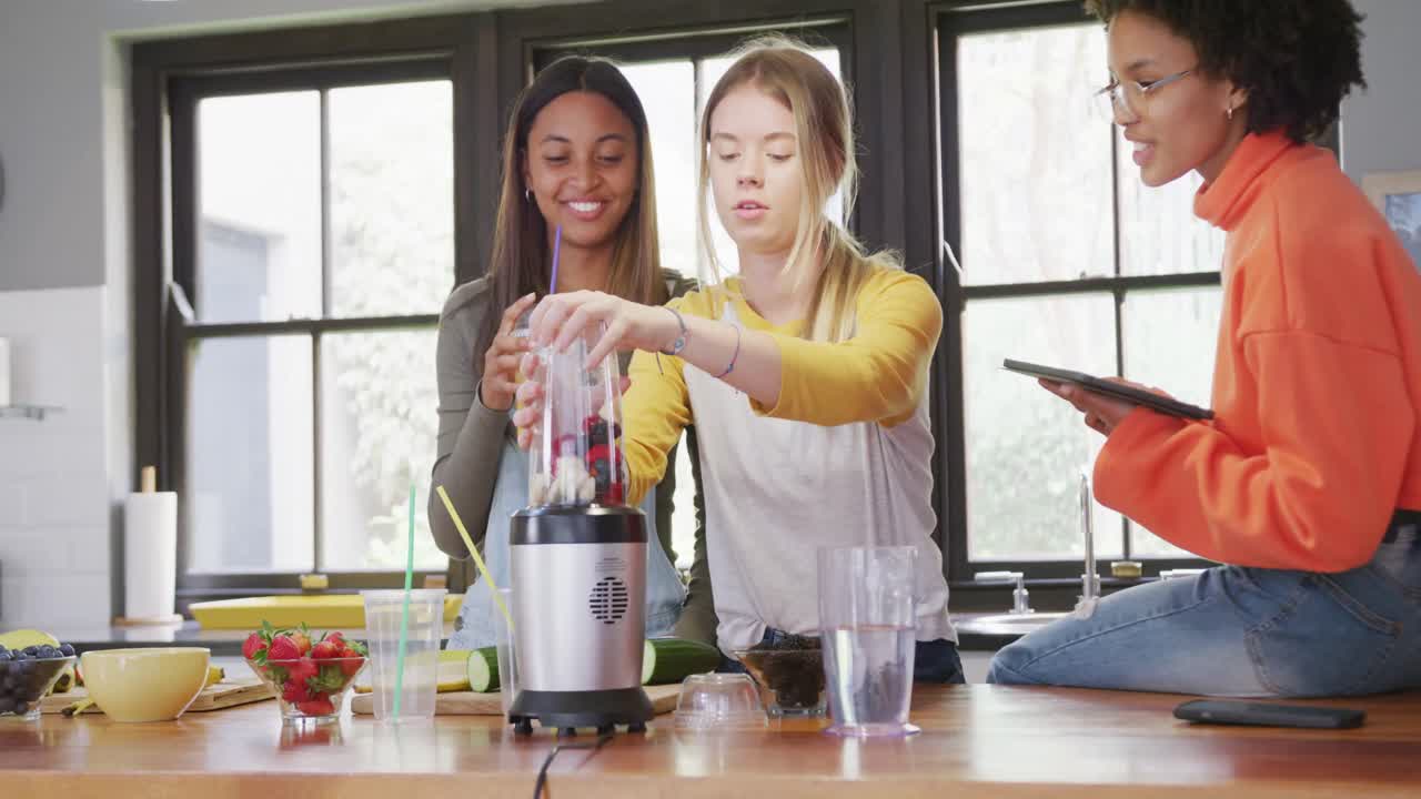 Amiche adolescenti felici di diverse origini che preparano una bevanda salutare usando un tablet in cucina, al rallentatore.