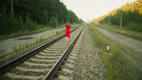 rear view of woman in red dress running along rail track while looking back, surrounded by forest vegetation with poles and rural structures visible in distance under soft natural daylight