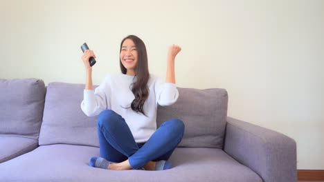 Young-asian-female-with-TV-remote-watching-sport-game-on-television,-celebrates-victory-of-her-team,-full-frame,-slow-motion