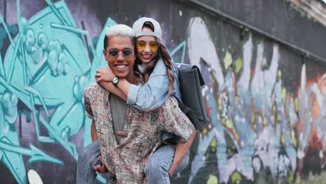 un joven que le da a su novia un piggyback y mira la cámara cerca de una pared de graffiti en la calle