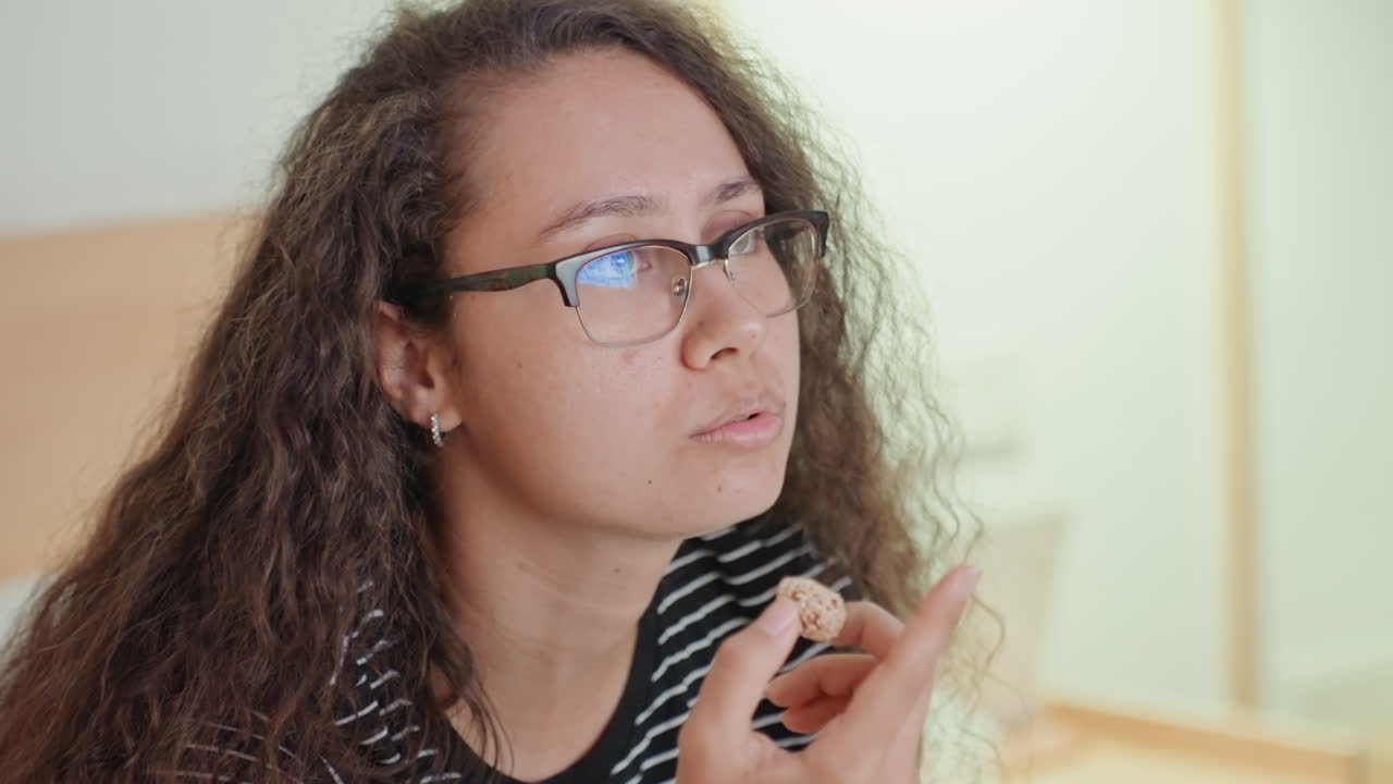 Ospite in camera d'albergo si prepara per una chiamata. Studente o lavoratore da remoto con maglietta a righe e occhiali contempla l'applicazione di balsamo o trucco mentre guarda verso la zona del laptop, letto e testiera visibili, concentrato.