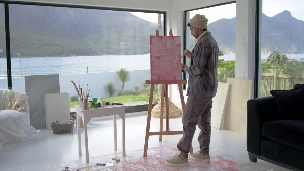 Painter is mixing paint in coastal studio, moving to easel and refining pink canvas for finishing