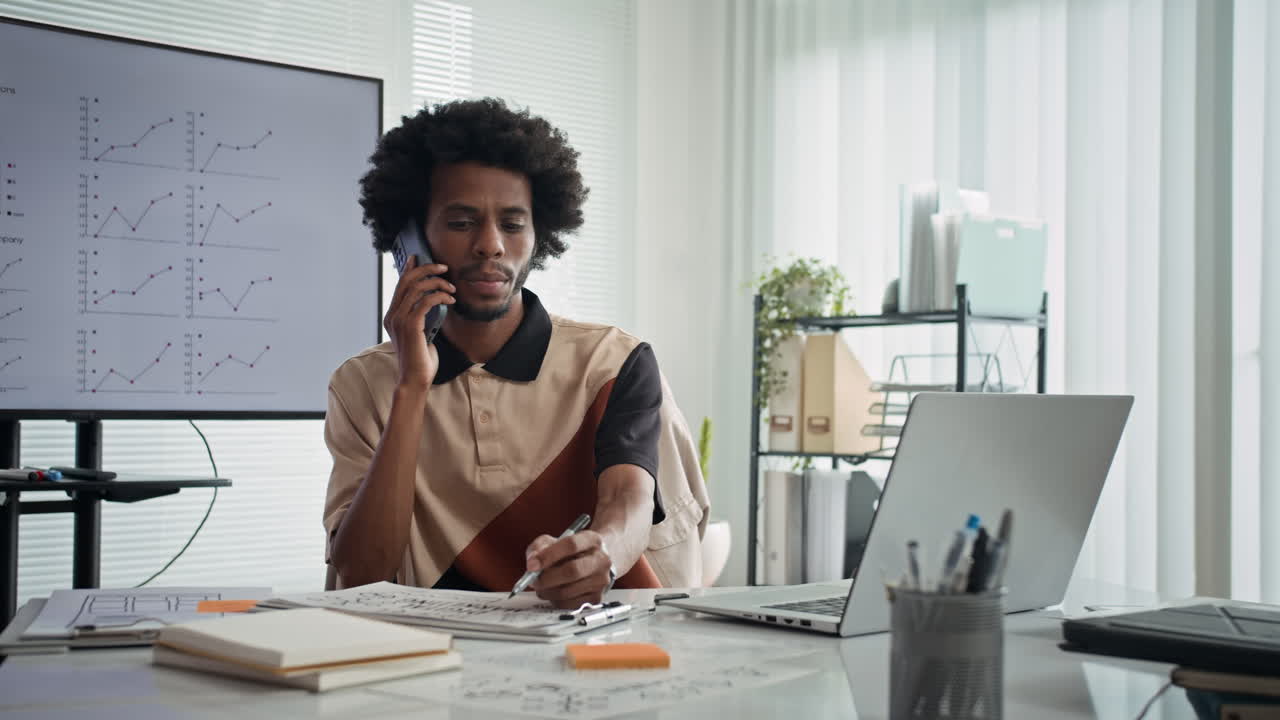 Sort administrator, der taler i smartphone ved en laptop på kontoret