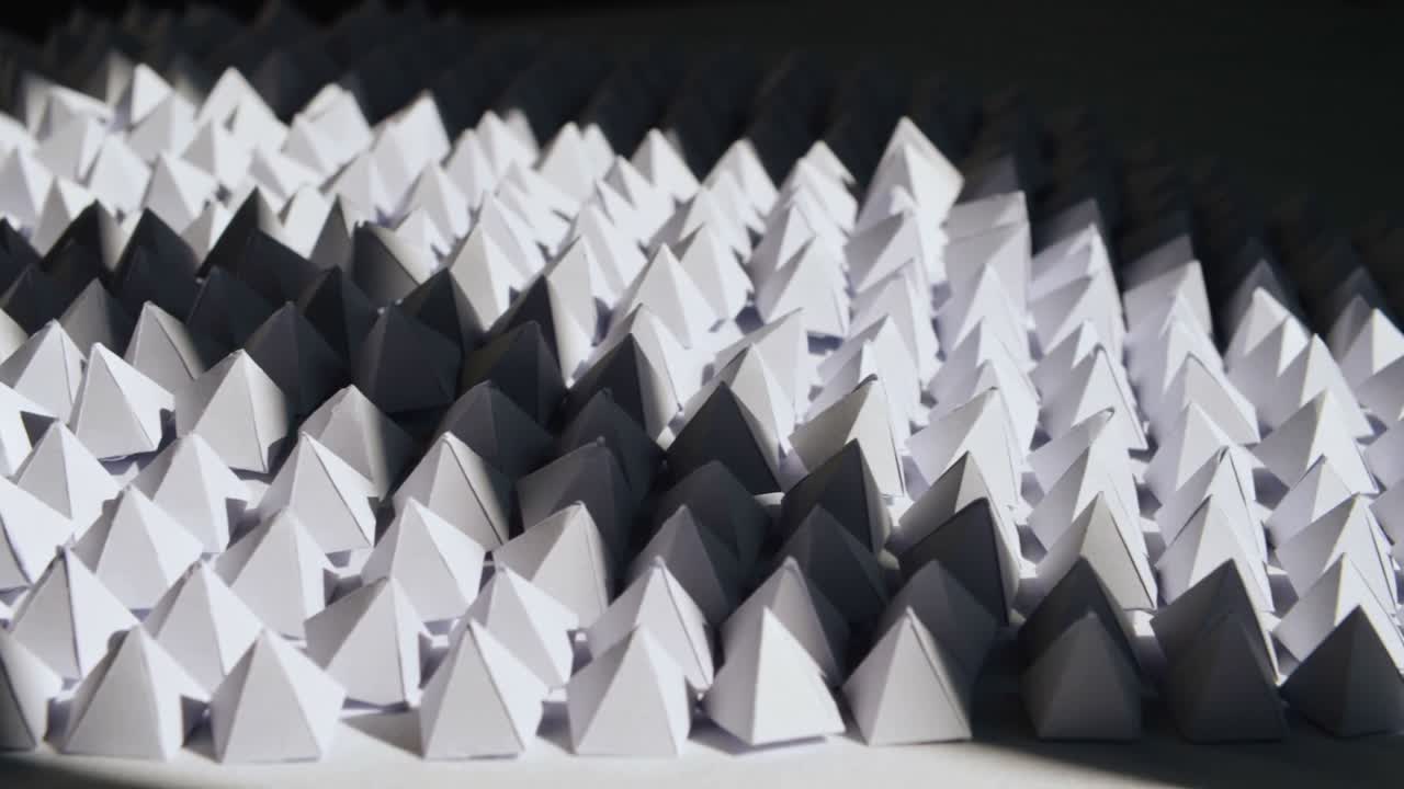 Panning camera revealing shifting shadows across grid of small white paper pyramids on tabletop