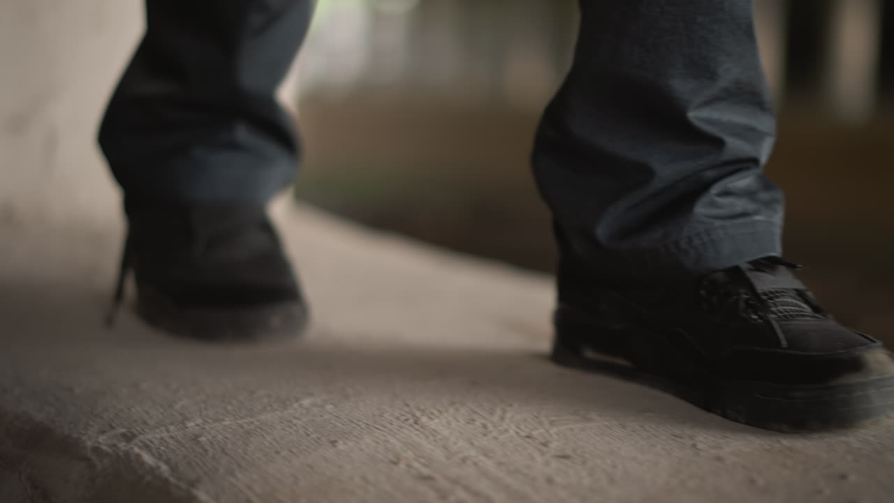 Primo piano di una scarpa da ginnastica bagnata che calpesta il cemento, gocce di pioggia al rallentatore che schizzano, luccichio di pozzanghere riflettenti sotto il bagliore di un sottopassaggio, risvolto in denim che sfiora la superficie granulosa, passo del pendolare che trasmette determinazione.