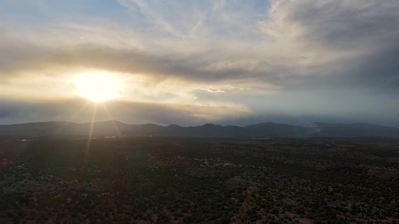 4K drone timelapse flies over rugged desert mesas toward a distant mountain sunset. Cinematic aerial motion captures bright sun rays piercing between two layers of dark, shifting stormy clouds.