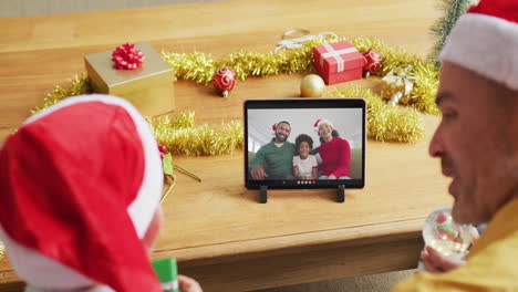 padre y hijo caucásicos con sombreros de santa usando una tableta para una videollamada de navidad con la familia en la pantalla