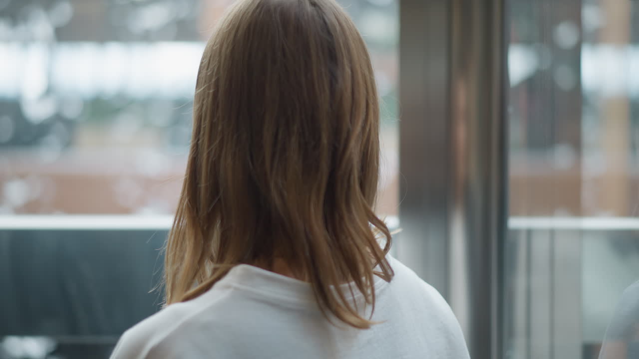 유리 패널과 흐릿한 자연 빛 반사를 통해 현대적인 도시 환경을 강조하는 부드러운 조명으로 움직이는 엘리베이터 내부의 젊은 여성의 뒷면