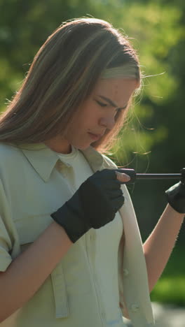 chica de pelo rubio de pie al aire libre con una expresión reflexiva, inspeccionando cuidadosamente el mango de la bomba de aire verde, el fondo suavemente borroso de los árboles y un edificio
