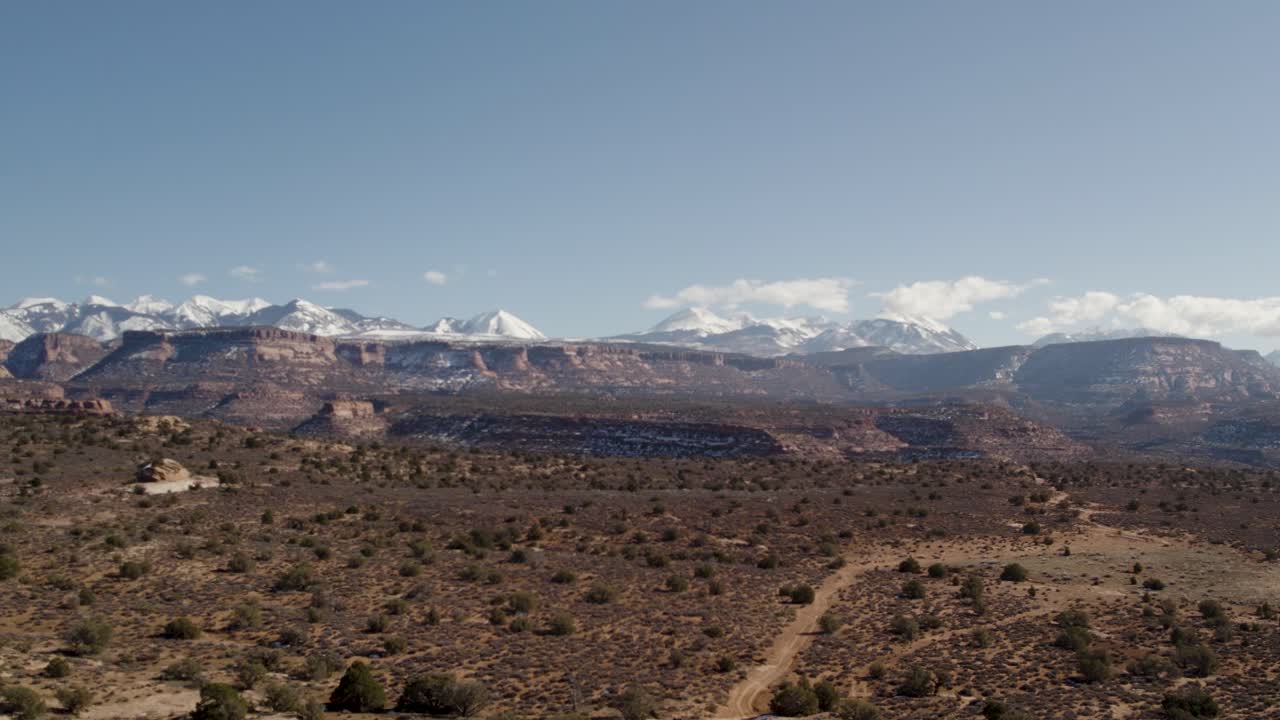 A high-flying drone shot over a remote dirt road cutting through the vast and unique desert land near Moab, Utah, with the snowy Rocky Mountains towering in the distance