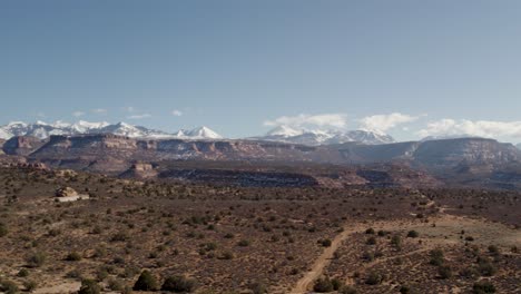 A-high-flying-drone-shot-over-a-remote-dirt-road-cutting-through-the-vast-and-unique-desert-land-near-Moab,-Utah,-with-the-snowy-Rocky-Mountains-towering-in-the-distance