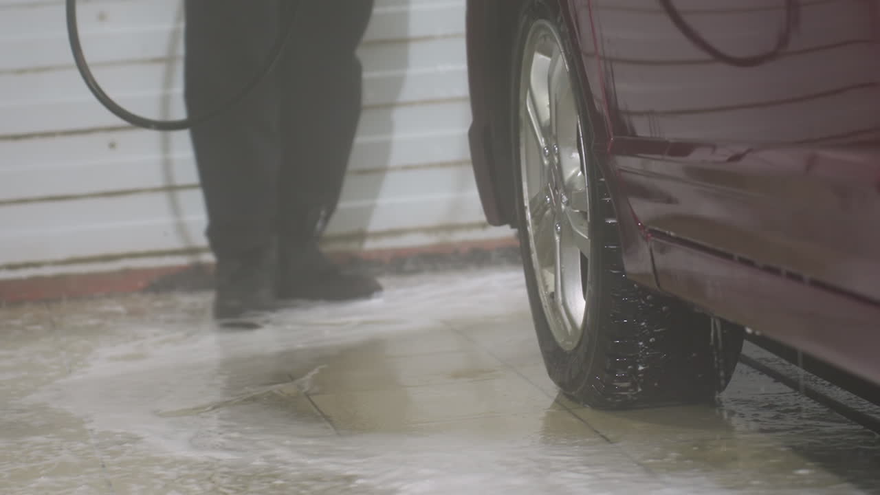 low angle close up of person wearing black pants and canvas shoes rinsing red car exterior with high pressure water spray inside indoor garage floor covered in soap and water puddles
